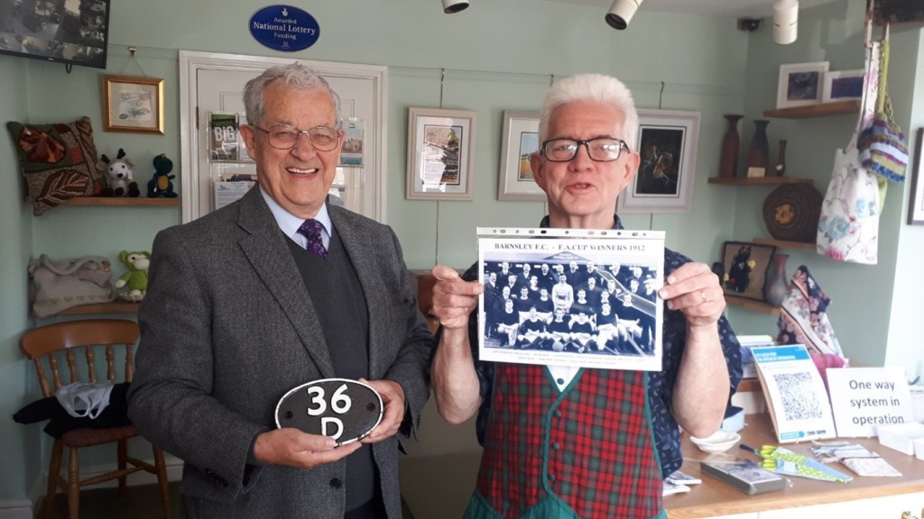Ken and Ian stood in the museum, they are holding objectys and photos behind them is the shop