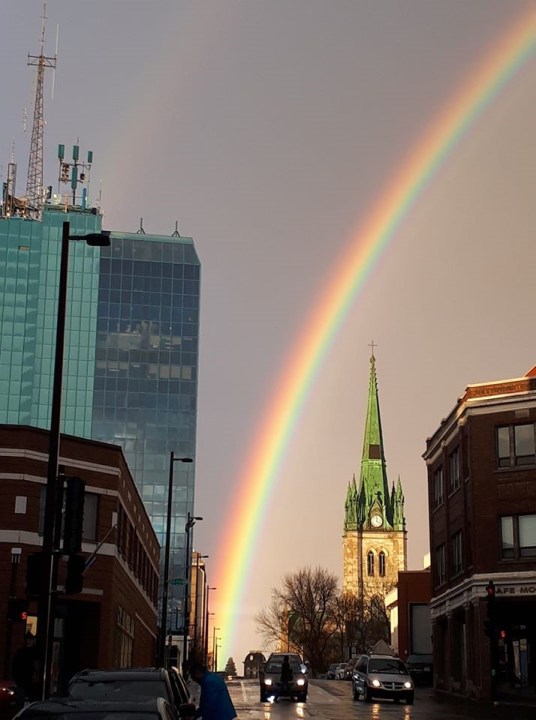 Title: Montreal Rainbow (For Leonard Cohen), an image by Josie Vie (c) 2022.
A vibrant rainbow falls over a hilly street in downtown Montreal, early 2th century buildings, an 18th century Cathedral tower, a modern glass skyscraper juxtaposed -- cars with headlights on waiting at a stoplight, front of cars facing the viewer.